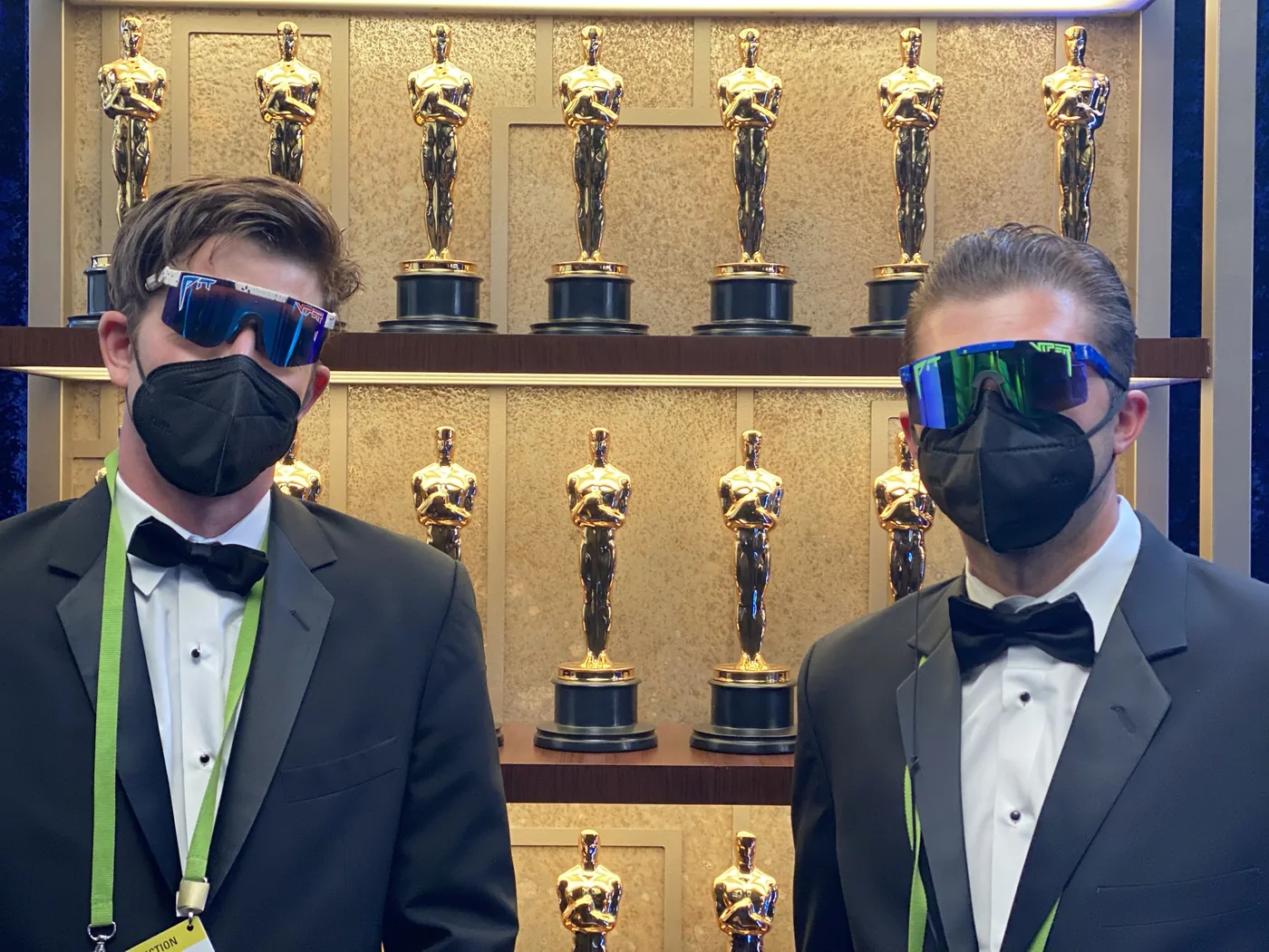 Two crew members in tuxedos backstage in front of shelves of Oscar statuettes