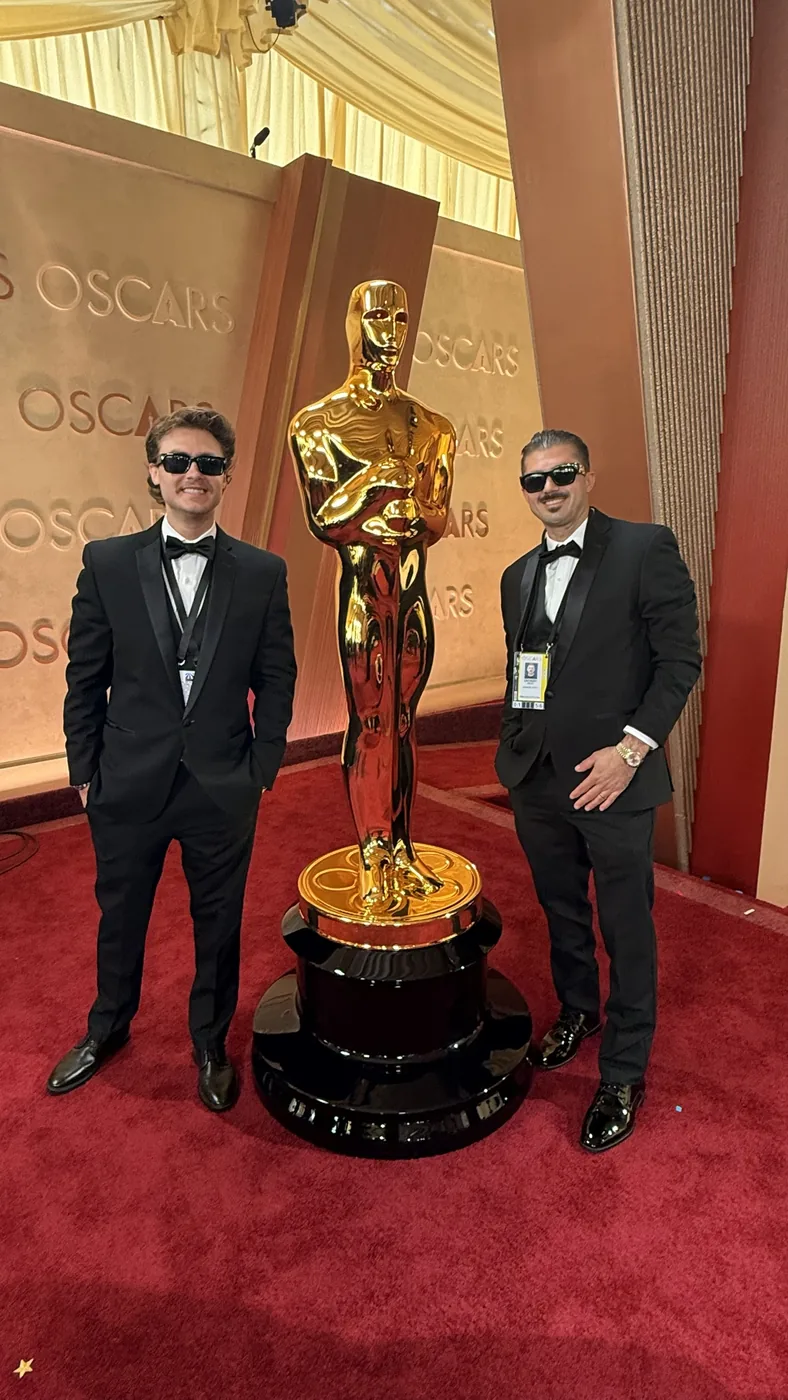 Two crew members in tuxedos on the Oscars red carpet beside the giant Oscar statue